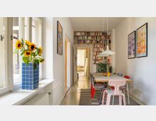 Ein heller Raum mit einer langen Tischgruppe aus verschiedenen bunten Stühlen und einer hängenden Deckenlampe. Links stehen große Fenster mit einer Vase und Sonnenblumen auf der Fensterbank, rechts befindet sich ein Bücherregal, dekorative Poster und eine Grünpflanze.