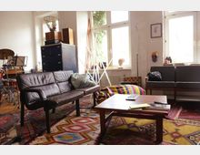 Wohnzimmer mit retro-inspirierter Einrichtung, bestehend aus einem braunen Ledersofa, einem niedrigen Holztisch und gemusterten Teppichen. Große Fenster lassen Tageslicht ein, während Möbel und Dekor wie ein Schlagzeug, bunte Kissen und eine Kommode das kreative Ambiente betonen.