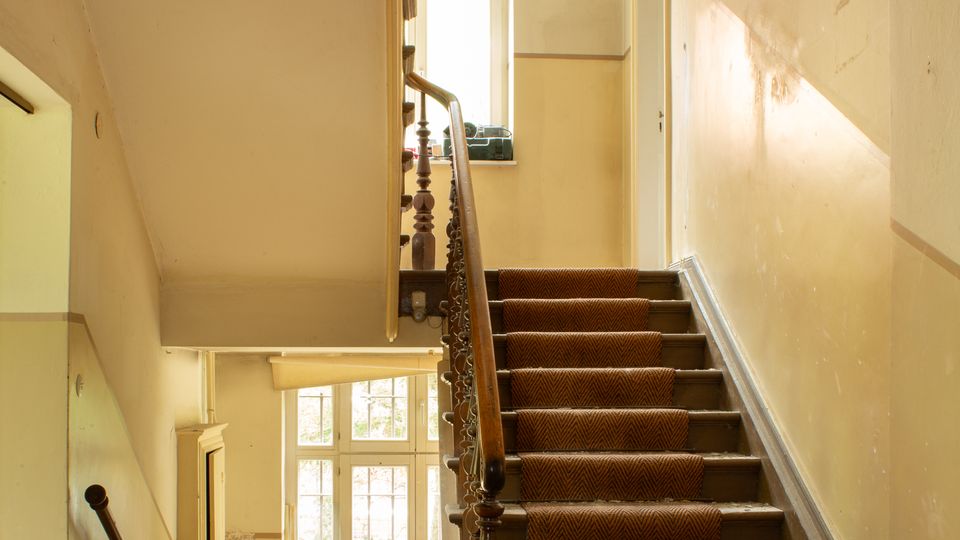 Treppenhaus in einem älteren, teils renovierungsbedürftigen Gebäude mit brauner Teppichläufer-Treppe, dunklem Holzgeländer und beigefarbenen Wänden. Große Fenster bringen Tageslicht herein; unten sind ein Heizkörper und eine Zwischenpodestfläche mit Staub und Bauschutt zu sehen.
