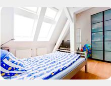 Helles Schlafzimmer im Dachgeschoss mit schrägen Decken, großen Dachfenstern und weißen Sichtbalken. Im Raum stehen ein Bett mit blau gemusterter Bettwäsche, ein großer Kleiderschrank mit Schiebetüren sowie ein kleiner TV-Bereich auf hellem Holzboden.