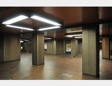 Unterirdischer Bahnhofsvorraum mit beigefarbenen Wänden, mehreren Säulen, einem gefliesten Boden in Brauntönen und rechteckigen Deckenleuchten. Im Hintergrund befindet sich ein gelber Fahrkartenautomat.