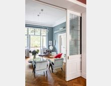 Wohnzimmer in einer Altbauwohnung mit hohen Decken, blau gestrichenen Wänden und Stuckverzierungen. Zu sehen sind ein Holzboden im Fischgrätmuster, eine Sitzecke mit modernen Sesseln und einem runden Glastisch, sowie große, bodentiefe Fenster mit Blick ins Grüne.