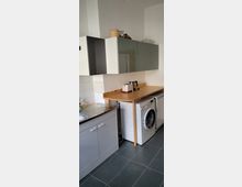 Eine kleine Küche mit grauen Bodenfliesen, einem Waschbecken mit Edelstahl-Abtropffläche, weißen und grauen Schränken sowie einer Arbeitsfläche aus Holz. Unter der Arbeitsfläche befinden sich eine Waschmaschine und ein Kühlschrank.