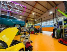 Innenraum einer Autowerkstatt mit orangefarbenem Boden, hohen Deckenbalken und großen Rolltoren mit Fenstern. Zu sehen sind ein Oldtimer auf einer Hebebühne, ein gelbes Auto mit geöffneter Motorhaube sowie zahlreiche Werkzeuge, Regale und Reifen; an der Decke hängen geometrische Neonleuchten.