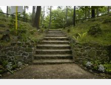 Steinerne Treppe in einem bewaldeten Park, umgeben von niedrigen Mauern aus Natursteinen, moosbewachsenen Böschungen und Pflanzen wie Farn und Blumen. Im Hintergrund sind Bäume und dichtes Grün zu sehen.