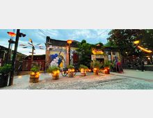 Außenaufnahme eines gepflasterten Hofs mit beleuchteten Pflanzenkübeln, Wandgemälden und einem flachen Ziegelgebäude. Im Hintergrund befinden sich Lichterketten, ein Kunstobjekt und Bäume; die Szenerie wirkt urban und kreativ.