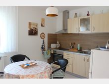 Helle Küche mit einer Küchenzeile aus hellem Holz, einer Arbeitsplatte und einem eingebauten Herd. Ein runder Tisch mit gemusterter Tischdecke und zwei Stühlen steht im Vordergrund, während an den Wänden Bilder und Dekorationen hängen.