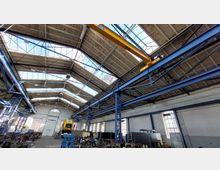 Industriehalle mit hohen Decken und großen Dachfenstern, die viel Tageslicht hereinlassen. Die Halle enthält Metallrahmenstrukturen, eine gelbe Laufkranbahn und industrielle Geräte sowie Arbeitsbereiche im unteren Bereich.