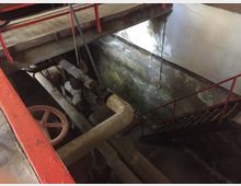 Industrieanlage mit verrosteten Rohren, einem großen roten Ventil und roten Metallgeländern. Im Hintergrund ist eine Betontreppe sichtbar, die hinunter in einen feuchten Raum mit fleckiger, abgenutzter Wand führt.