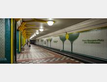 U-Bahn-Station mit großzügigem, langgezogenem Bahnsteig, dekoriert mit gekachelten Wänden, die Baummotive und den Schriftzug „Franz-Neumann-Platz – Am Schäfersee“ zeigen. Gelbe und grüne Säulen sowie leuchtende Deckenlampen prägen die Gestaltung; der Boden besteht aus gemusterten Fliesen.