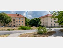 Zwei gegenüberliegende Wohngebäude mit beige-farbenen Fassaden und roten Ziegeldächern, dazwischen ein kleiner Kreisverkehr mit Grünflächen und einem großen Baum. Im Vordergrund ein gepflasterter Gehweg und vereinzelte Büsche.