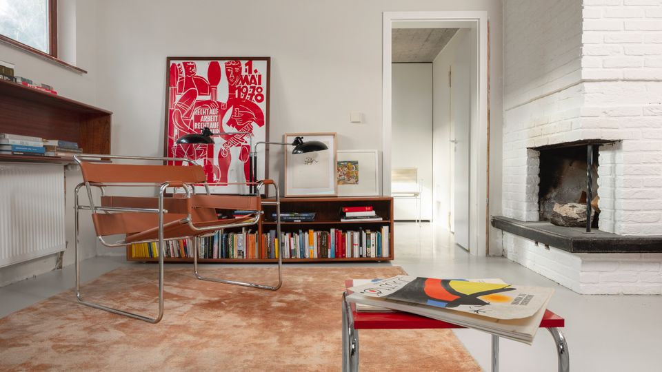 Modern minimalist living room with a raw concrete ceiling, white walls, and a white brick fireplace with a dark hearth. A low bookcase with framed art sits against the wall beside chrome-framed chairs on a rug, with a large window and doorway leading to another room.