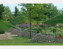 Ein Landschaftsgarten mit blühenden Beeten, gepflasterten Wegen und niedrigen Steinmauern, umgeben von grünen Grashügeln und Bäumen. Im Hintergrund sind schmale Treppen und ein Gebäude mit Flachdach zu erkennen.