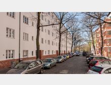 Wohnstraße mit mehrstöckigen Wohngebäuden aus hellem Putz und roten Ziegeln im unteren Bereich. Die Straße ist gepflastert, gesäumt von parkenden Autos und Bäumen ohne Laub.