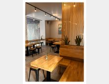 Ein modernes Café mit Holztischen, Bänken und schwarzen Metallstühlen. Im Hintergrund sind eine gemusterte Wand, ein dekoratives Gemälde und eine Raumtrennung aus vertikalen Holzlamellen sichtbar, ergänzt durch Zimmerpflanzen.