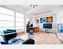 Heller Wohnbereich in einer Dachgeschosswohnung mit großen Fensterfronten, hellem Holzfußboden und weißen, leicht schrägen Deckenbalken. Links stehen Sofa und Sessel, mittig ein kleiner Glastisch, rechts ein Fernseher mit Lowboard und Wandbildern darüber sowie ein Arbeitsbereich mit Tisch.