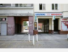 Fassade eines Wohn- und Geschäftshauses mit zwei Durchgängen, die zu Hinterhöfen führen. Links ist eine türkisfarbene Wand mit Graffiti, rechts ein Laden mit einem „Grundig“-Schild, davor stehen weiße Poller auf einem gepflasterten Gehweg.