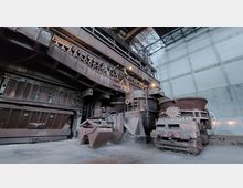 Industriehalle mit großer, rostiger Stahlkonstruktion und mehreren massiven Schmelzkesseln. Die Halle hat hohe, weiße Wände, ein Gitterdach und wirkt wie ein ehemaliger Bereich eines Stahlwerks oder einer Gießerei.