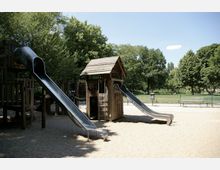 Ein Kinderspielplatz mit Holzspielgeräten, darunter zwei Metallrutschen, ein Kletterbereich und ein kleines Häuschen mit Leiter, umgeben von Sandboden. Im Hintergrund sind Bäume, eine Wiese und Sitzbänke zu sehen.
