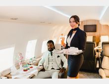 Ein luxuriöser Privatjet-Innenraum mit eleganten Ledersitzen und gedeckten Tischen. Eine Flugbegleiterin in Uniform serviert einem Passagier, der in einem bequemen Sitz Platz genommen hat, ein Getränk und eine Mahlzeit.