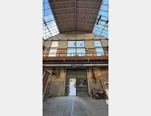 Innenansicht einer alten Industriehalle mit hohem Holzdach, Stahlträgern und großen Rasterfenstern entlang der Backsteinwand. Unten befindet sich ein großes, teilweise geöffnetes Metalltor mit sichtbaren Rohren und verstreuten Werkstattmaterialien am Rand.