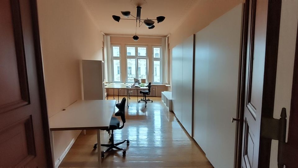 Langer, schmaler Büroraum mit Holzboden, mehreren Schreibtischen und Rollstühlen sowie hohen Einbauschränken an der rechten Wand. Am Ende des Raums befindet sich ein großes Fenster mit Blick nach draußen, darüber eine moderne Deckenleuchte.