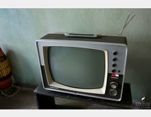 Ein älterer Röhrenfernseher mit grauem Gehäuse, abgerundeter Bildschirmfläche und Drehknöpfen an der Vorderseite steht auf einem schwarzen Tisch vor einer grünlichen Wand. Antennen sind an der Rückseite angebracht.