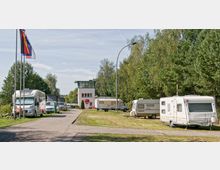 Wohnmobilstellplatz mit mehreren geparkten Wohnmobilen und Wohnwagen, umgeben von grünen Bäumen. Im Hintergrund steht ein kleines, weißes Gebäude mit Flachdach und grünen Fensterrahmen, daneben wehen Flaggen an Fahnenmasten.