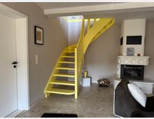 Innenraum eines Wohnzimmers mit einer gelben Wendeltreppe, die zu einer oberen Etage führt. Rechts steht ein weißer Kachelkamin mit Holzstapel daneben, links befindet sich eine weiße Tür, und der Raum ist mit hellen Wänden und einem steinfarbenen Boden ausgestattet.