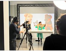 Ein Film- oder Fotostudio mit Beleuchtung, Kameras und einem Team bei der Arbeit. Im Vordergrund filmt eine Person mit einer Kamera, während eine andere Person einen Hund auf einem Tisch präsentiert und ein Hintergrund mit Bildern von Tieren beleuchtet ist.