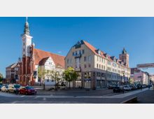 Städtisches Straßenzentrum mit einer Kirche und einer Reihe von Wohn- und Geschäftshäusern. Die Kirche hat einen weißen Turm mit grüner Spitze und roten Backsteingiebeln, daneben moderne Gebäude mit roten Dächern, parkende Autos und ein Straßenschild mit der Aufschrift „Schmalzgasse“.