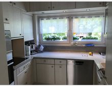 Helle Küche mit weißen Holzschränken, einem grauen Arbeitsplattenbereich und einem großen Fenster mit Blick ins Grüne. Auf der Arbeitsfläche stehen Haushaltsgeräte, eine Brille sowie kleinere Dekorationsgegenstände.