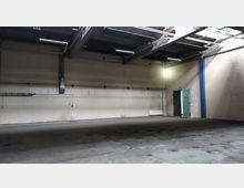 Leere Industriehalle mit hohen Decken, sichtbaren Stahlträgern und Rohren. Die Wände sind beige gestrichen, und es gibt eine geöffnete grüne Stahltür sowie einfache Beleuchtungskörper an der Decke.