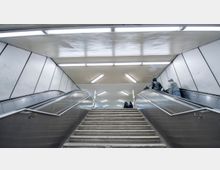 Treppenaufgang und Rolltreppen in einer U-Bahn-Station mit weißen Wänden und Deckenleuchten; Personen nutzen die Rolltreppen auf beiden Seiten.