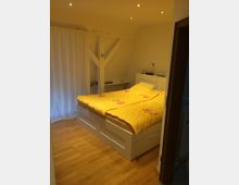 Schlafzimmer mit Doppelbett in weißem Rahmen und gelber Bettwäsche, Holzboden und Spotbeleuchtung an der Decke. Im Hintergrund sind ein weißer Vorhang, ein kleiner Nachttisch und eine sichtbare Deckenbalkenkonstruktion zu sehen.