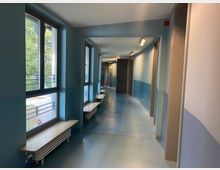 Ein langer, lichtdurchfluteter Flur mit hellblauen Wänden und einem blauen Boden. Auf der linken Seite befinden sich große Fenster mit darunterliegenden Sitzbänken, die Heizkörper verdecken, während der Flur am Ende in eine Tür mündet.