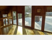 Innenraum eines Holzhauses mit großen Fenstern, die auf eine verschneite Außenlandschaft blicken. Das Sonnenlicht fällt durch die Fenster auf den hellen Holzboden, und die Wände sind komplett aus Holzpaneelen gefertigt.