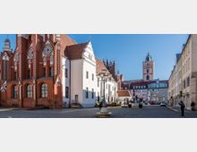 Blick auf einen historischen Platz mit Gebäuden in verschiedenen Architekturstilen, darunter eine rote Backsteinkirche mit gotischen Fenstern und ein weißes Gebäude mit Satteldach. Im Hintergrund ragt ein Kirchturm mit Uhr hervor, umgeben von modernen und traditionellen Fassaden; Straßenlaternen und Sitzbereiche prägen die offene Fläche.
