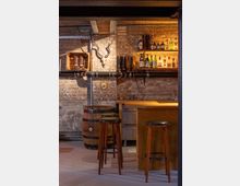 Rustikale Bar mit Backsteinwänden, einem hölzernen Tresen und hohen Barhockern. Im Hintergrund stehen Regalflächen mit Spirituosen, Cowboy-Stiefeln und ein Geweih an der Wand, ergänzt durch ein dekoratives Holzfass.