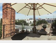 Terrasse mit Holzmöbeln und Topfpflanzen, teilweise beschattet durch einen großen Sonnenschirm; im Hintergrund ein roter Ziegelkamin, moderne Stadtgebäude und der Berliner Fernsehturm sind sichtbar.