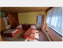 Zimmer mit zwei Einzelbetten mit rot-gemusterten Bezügen, einer Kommode dazwischen, gelb gestrichener Wand, Holzdecke und einem großen Fenster mit weißen Vorhängen. Rechts führt eine geöffnete Tür zu einem angrenzenden Raum mit Fliesenboden.