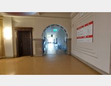 Ein heller Flur in einem historischen Gebäude mit gewölbtem Durchgang, Holztür auf der linken Seite, und Wandbeschilderung mit roten Markierungen rechts. Am Ende des Flurs ist ein grünes Notausgangsschild und Tageslicht sichtbar.