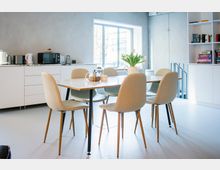 Heller, moderner Küchen- und Essbereich mit großem Fenster, weißer Küchenzeile mit Elektrogeräten und einem Esstisch mit sechs gepolsterten Stühlen. Rechts steht ein offenes Regal mit Büchern und Spielen; auf dem Tisch stehen Teekanne und eine Vase mit Blumen.