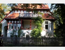 Zweistöckiges Wohnhaus mit rotem Ziegeldach, dekorativen Fachwerkelementen und einem Balkon. Die Fassade ist teilweise mit Rankpflanzen bewachsen, und ein Holzzaun umgibt das Grundstück; im Hintergrund stehen Bäume in einem Garten.
