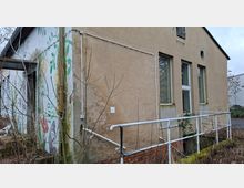 Seitenansicht eines älteren, verputzten Gebäudes mit drei hohen Fenstern und einer kleinen Rampe mit weißem Metallgeländer entlang der Fassade. Links ist ein Wandgemälde mit Pflanzenmotiven zu sehen; davor stehen kahle Sträucher und überwucherte Bereiche am Sockel aus Ziegeln.