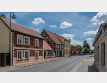 Straßenszene in einer kleinen Stadt mit historischen Häusern, darunter Fachwerk- und Backsteingebäude mit roten Ziegeldächern. Die Straße ist asphaltiert und von einem Bürgersteig mit Straßenlaternen gesäumt, im Hintergrund stehen Bäume unter einem blauen Himmel mit wenigen Wolken.