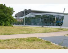 Moderne Sporthalle mit geschwungenem Dach und großer Glasfassade, umgeben von Grünflächen und Wegen. Vor dem Gebäude befinden sich Fahrradständer sowie eine offene Fläche für Fußgänger.