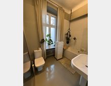 Bright bathroom with a large frosted window framed by beige curtains, a wall-mounted toilet, and a freestanding white sink. A built-in bathtub with light tile surround sits along the right wall, with a hanging plant and a slim wheeled storage cabinet beside a radiator under the window.