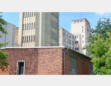 Backsteinbau mit einem flachen Dach im Vordergrund und höheren Gebäuden aus Beton mit schmalen Fenstern im Hintergrund. Umgeben von Bäumen und strahlend blauem Himmel.