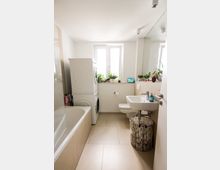 Helles Badezimmer mit beigefarbenen Fliesen, Badewanne links, wandhängendem Waschbecken und Toilette. Ein stapelbarer Wasch- und Trockner steht vor einem Fenster, das von mehreren Topfpflanzen dekoriert ist.